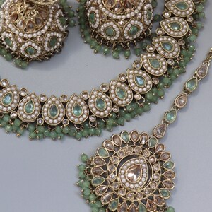 Op de afbeelding: Een gouden ketting en oorbellen set met groene en witte stenen. De ketting heeft een traanvormig ontwerp en de oorbellen zijn rond.