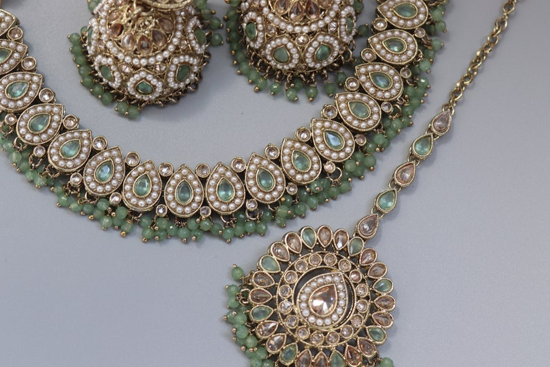 Op de afbeelding: Een gouden ketting en oorbellen set met groene en witte stenen. De ketting heeft een traanvormig ontwerp en de oorbellen zijn rond.