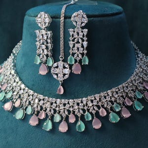 May include: A silver-toned jewelry set featuring a necklace, earrings, and a headpiece. The set is adorned with clear crystals and teardrop-shaped pink and green gemstones. The jewelry is displayed on a teal-colored surface.