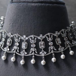Könnte beinhalten: Ein silberfarbenes Choker-Halsband mit filigranem Design. Die Halskette hat klare, rechteckige und runde Steine mit hängenden weißen Perlen. Die Halskette wird auf einem grauen Schaufensterkopf präsentiert.