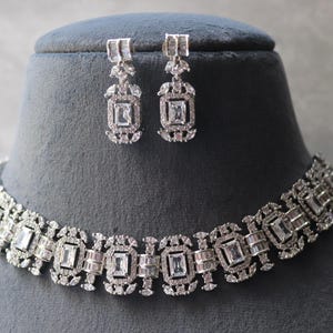 May include: A silver-toned necklace and matching earrings set. The jewelry features rectangular and square-cut clear stones set in an ornate design. The necklace is displayed on a gray mannequin head.