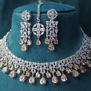 Pode incluir: Um conjunto de joias deslumbrante com colar, brincos e um adorno de cabeça. O conjunto é adornado com pedras preciosas transparentes e champanhe, cravejadas em metal prateado. O colar e os brincos têm um design em cascata, e o adorno de cabeça tem um pingente central.