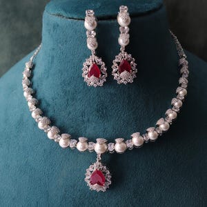 May include: A silver-toned necklace and earring set with a pearlescent and gemstone design. The necklace features a central pendant with a red gemstone, and the earrings have a matching design. The jewelry is displayed on a teal mannequin.
