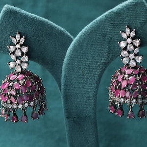 May include: A pair of dark metal jhumka earrings. The earrings have a floral design at the top with white stones, and a bell-shaped body adorned with ruby-coloured stones. Small ruby-coloured teardrops dangle from the bottom.