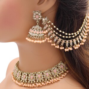 Könnte beinhalten: Ein peachfarbener Choker-Halskette und Ohrring-Set mit goldenen Akzenten und Perlendetails. Die Halskette und Ohrringe haben ein florales Design mit rosa und klaren Steinen.