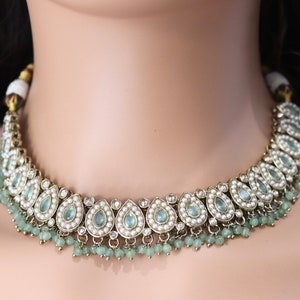 Op de afbeelding: Een zilveren en gouden ketting met traanvormige blauwe stenen en witte parels. De ketting heeft een rij groene kralen die aan de onderkant hangen.