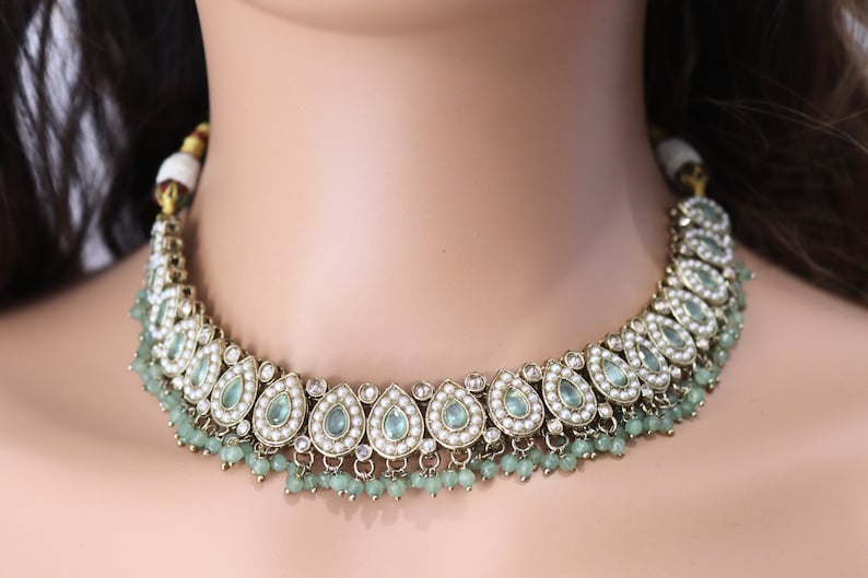Op de afbeelding: Een zilveren en gouden ketting met traanvormige blauwe stenen en witte parels. De ketting heeft een rij groene kralen die aan de onderkant hangen.