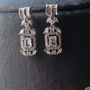 May include: A pair of silver-toned earrings with a rectangular central stone and smaller surrounding stones. The earrings have a rectangular top section and a decorative, geometric design. The earrings are displayed on a dark gray surface.