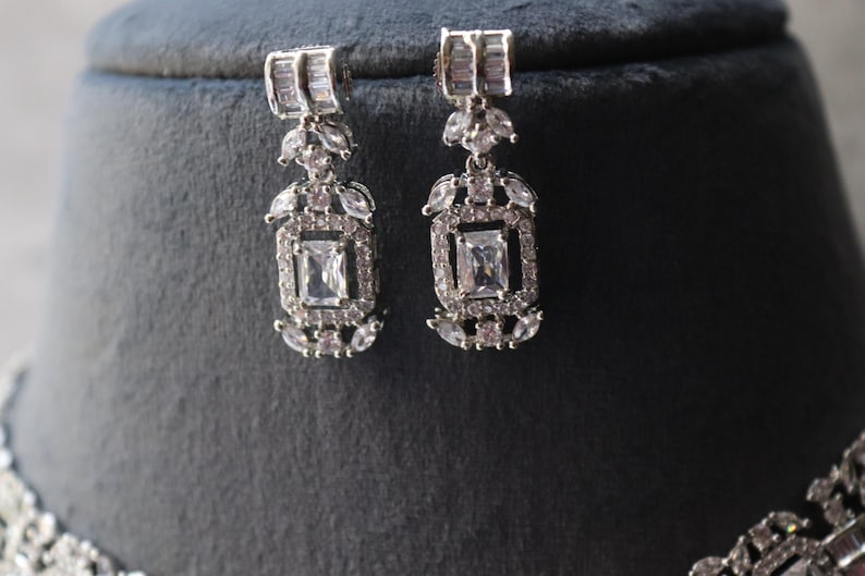 May include: A pair of silver-toned earrings with a rectangular central stone and smaller surrounding stones. The earrings have a rectangular top section and a decorative, geometric design. The earrings are displayed on a dark gray surface.