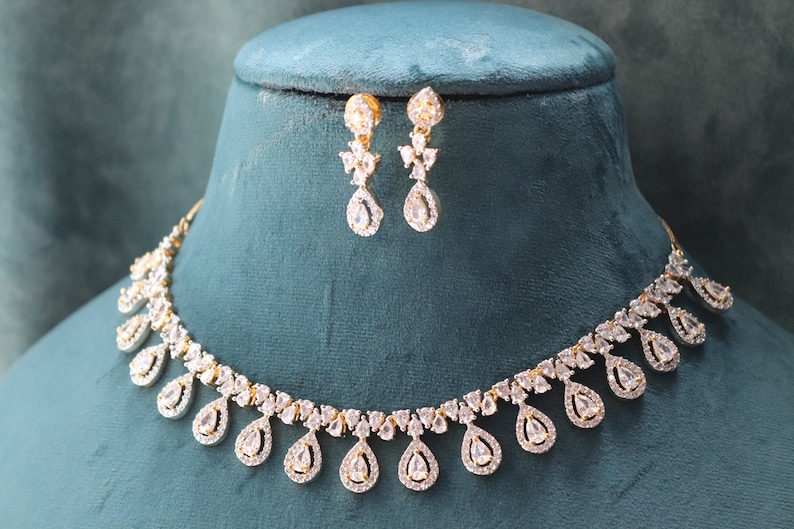 May include: A gold-toned necklace and matching earrings set. The jewelry features a series of teardrop-shaped, clear gemstones. The necklace and earrings are displayed on a teal-colored bust, showcasing the intricate design and sparkle.