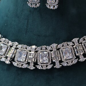 May include: A silver-toned necklace and earring set. The necklace features a series of large, square-cut, clear gemstones, interspersed with smaller, rectangular and oval-shaped stones. The earrings mirror the design. The jewelry is set against a dark green background.