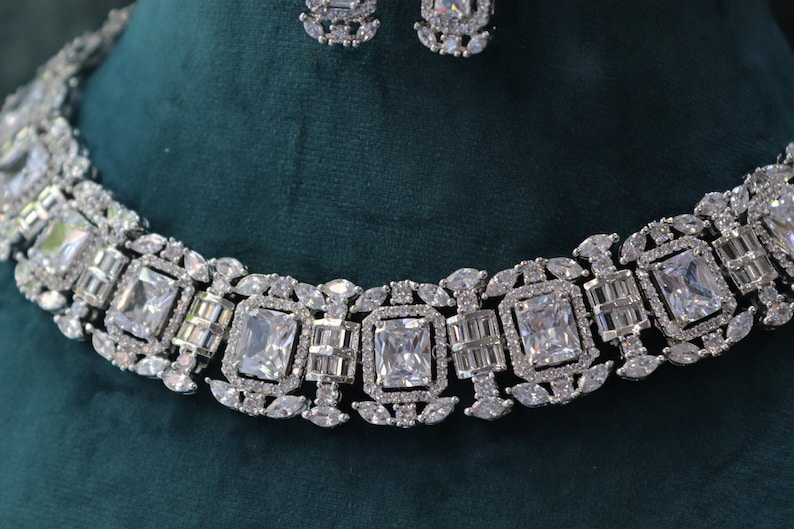 May include: A silver-toned necklace and earring set. The necklace features a series of large, square-cut, clear gemstones, interspersed with smaller, rectangular and oval-shaped stones. The earrings mirror the design. The jewelry is set against a dark green background.