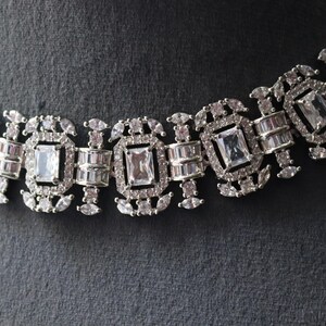 May include: Close-up of a silver-toned necklace with a repeating pattern of rectangular and oval-shaped clear gemstones. The necklace is set against a dark gray background, highlighting the intricate design and sparkle of the jewelry.