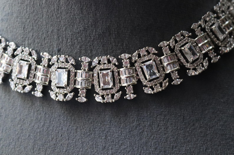 May include: Close-up of a silver-toned necklace with a repeating pattern of rectangular and oval-shaped clear gemstones. The necklace is set against a dark gray background, highlighting the intricate design and sparkle of the jewelry.