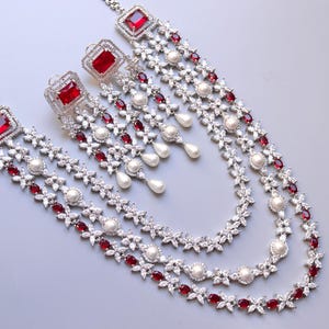May include: A silver-toned necklace and matching earrings set. The necklace features three strands of white and red gemstones, with a square red gemstone at the top. The earrings have a square red gemstone and dangling white pearls.