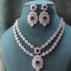 May include: A dazzling jewelry set featuring a two-strand necklace and matching earrings. The necklace and earrings are adorned with clear, sparkling stones and a central square, deep blue stone. The set is displayed on a teal mannequin.