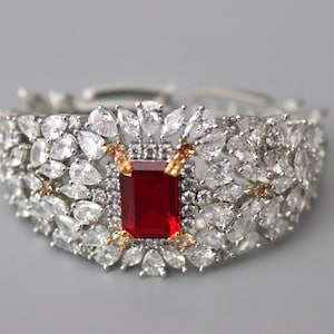 May include: A silver-toned bracelet with a central rectangular red gemstone, surrounded by numerous clear, sparkling crystals. The bracelet features a floral design with gold-toned accents, set against a gray background.