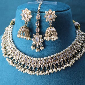 May include: A gold-coloured jewellery set comprising a necklace, earrings, and a headpiece. The necklace and earrings feature clear, oval-shaped stones and small white pearls. The headpiece has a floral design with dangling teardrop-shaped stones.