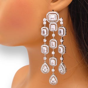 May include: Silver dangle earrings with multiple rows of clear cubic zirconia stones. The stones are in a square shape and are set in a silver frame. The earrings are long and have a teardrop shape at the bottom.