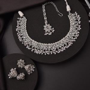 May include: A silver-toned jewelry set consisting of a necklace, a pair of jhumka earrings, and a maang tikka. Each piece features clear, teardrop-shaped stones arranged in floral patterns, accented with small white beads along the edges.
