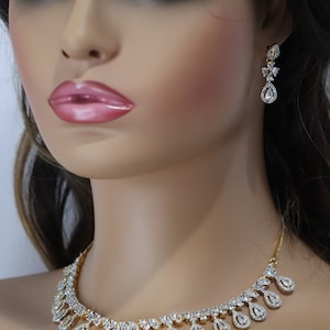May include: A gold-toned necklace and matching earrings set. The jewelry features a series of clear, sparkling stones set in a teardrop design. The necklace and earrings are displayed on a mannequin.