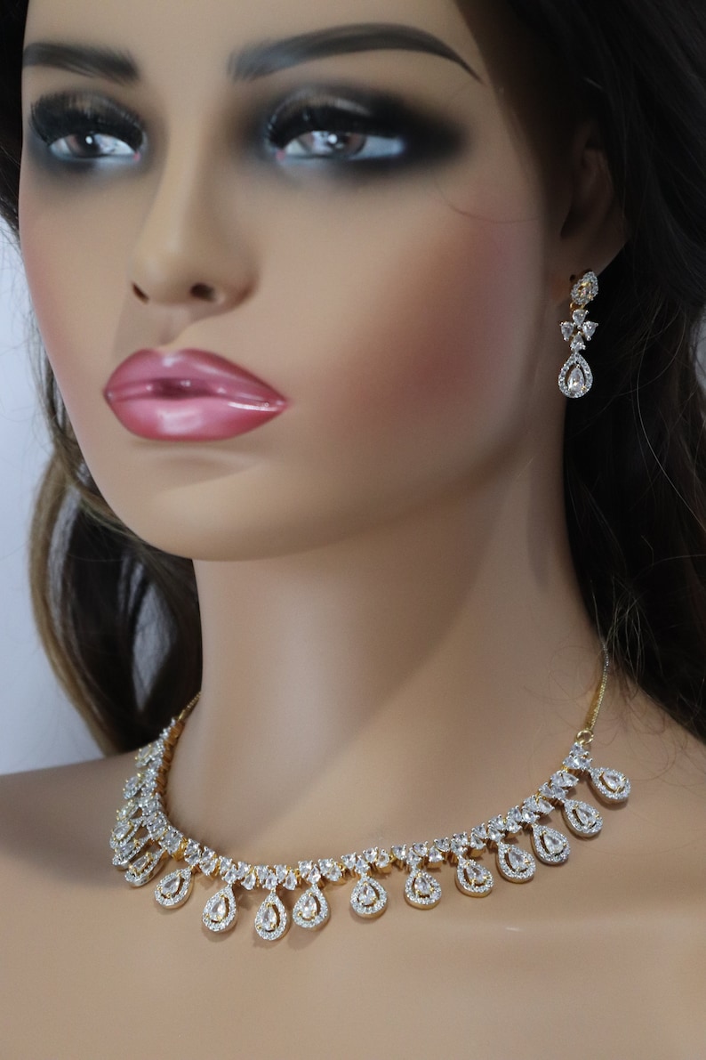 May include: A gold-toned necklace and matching earrings set. The jewelry features a series of clear, sparkling stones set in a teardrop design. The necklace and earrings are displayed on a mannequin.