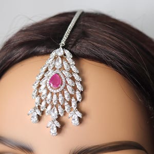 May include: A silver-toned headpiece with a central teardrop-shaped, pink gemstone, surrounded by clear, sparkling crystals. The headpiece is designed with a chain that sits atop the head, and the decorative portion dangles over the forehead.