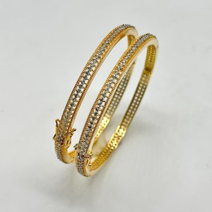 May include: Two gold-colored bangles with a repeating pattern of small, clear stones. The bangles have a hinged clasp and are displayed on a white surface. The jewelry is likely made of metal and is designed to be worn on the wrist.