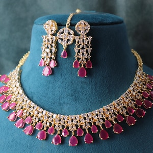 May include: A gold-coloured necklace, earrings, and headpiece set. The jewellery features clear, sparkling stones and teardrop-shaped, magenta-coloured gems. The set is displayed on a blue velvet bust.