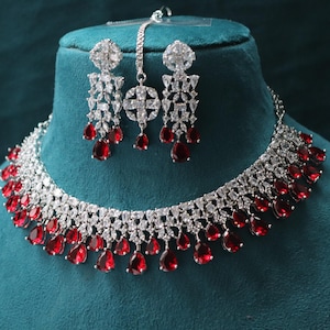 May include: A silver necklace and earrings set with a white stone design and red teardrop accents. The jewelry is displayed on a blue velvet stand.