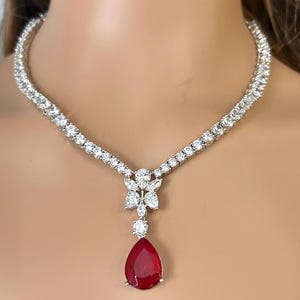 Peut inclure: Un collier étincelant orné de pierres précieuses rondes et transparentes serties dans un métal argenté. Le collier comporte un pendentif avec une grosse pierre précieuse rouge foncé en forme de goutte d'eau. Le pendentif est orné de petites pierres précieuses transparentes.
