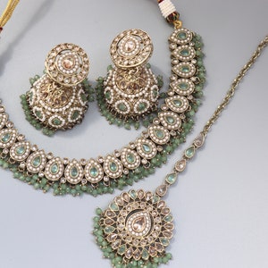 Op de afbeelding: Een Indiase bruidsjuwelenset in groen en goud, bestaande uit een ketting, oorbellen en een maang tikka. De ketting en oorbellen hebben druppelvormige stenen en parels, terwijl de maang tikka een rond ontwerp heeft.