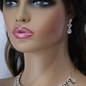 May include: A gold-toned necklace and matching earrings set. The necklace features a series of teardrop-shaped pendants, each encrusted with clear gemstones. The earrings have a similar design, with a teardrop pendant and smaller gemstones.