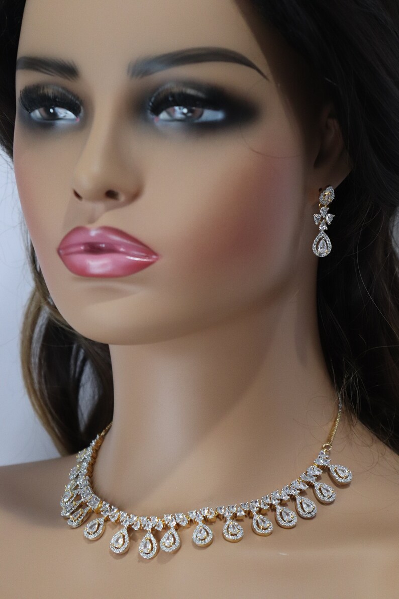 May include: A gold-toned necklace and matching earrings set. The necklace features a series of teardrop-shaped pendants, each encrusted with clear gemstones. The earrings have a similar design, with a teardrop pendant and smaller gemstones.