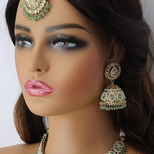 Op de afbeelding: Een Indiase bruidssieradenset in goud en groen met een ketting, oorbellen en een tikka. De ketting heeft een delicate ketting met een hanger van druppelvormige stenen en parels. De oorbellen zijn hangende oorbellen met een vergelijkbaar ontwerp als de ketting. De tikka is een traditionele voorhoofdversiering met een ronde hanger.