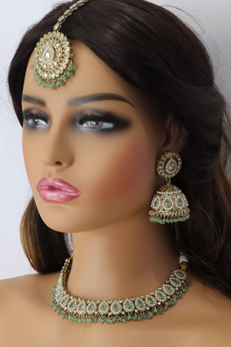 Op de afbeelding: Een Indiase bruidssieradenset in goud en groen met een ketting, oorbellen en een tikka. De ketting heeft een delicate ketting met een hanger van druppelvormige stenen en parels. De oorbellen zijn hangende oorbellen met een vergelijkbaar ontwerp als de ketting. De tikka is een traditionele voorhoofdversiering met een ronde hanger.