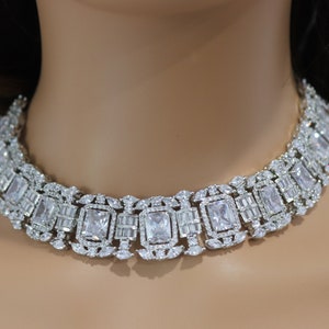 May include: A dazzling silver-toned necklace featuring a series of rectangular and baguette-cut clear crystals. The necklace is designed to sit around the neck, showcasing a luxurious and elegant style. The crystals are set in a detailed, ornate pattern.