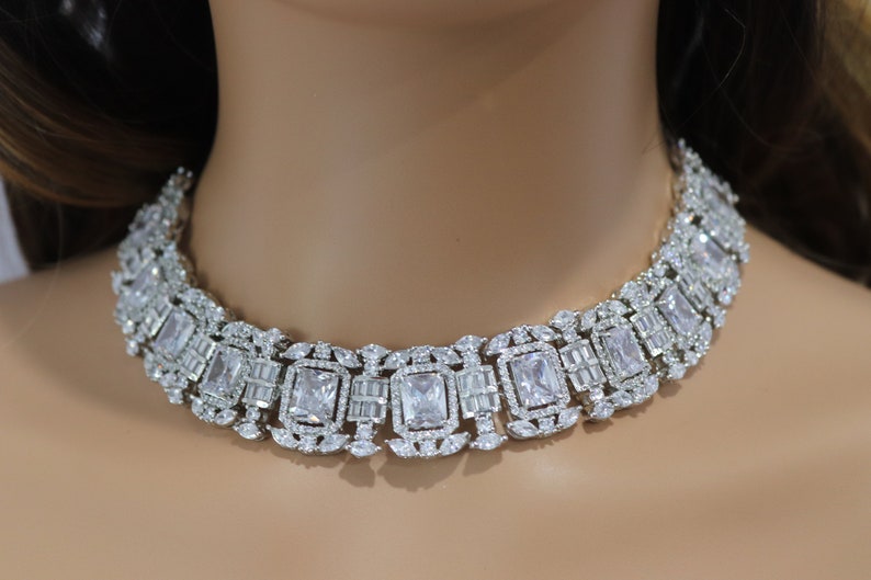 May include: A dazzling silver-toned necklace featuring a series of rectangular and baguette-cut clear crystals. The necklace is designed to sit around the neck, showcasing a luxurious and elegant style. The crystals are set in a detailed, ornate pattern.