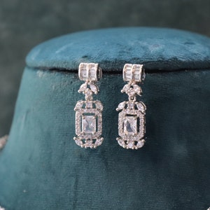 May include: Elegant silver-toned earrings with a rectangular, clear gemstone center and smaller surrounding stones. The earrings are displayed on a dark green velvet surface, showcasing their intricate design and sparkle. The earrings are part of a jewelry set.