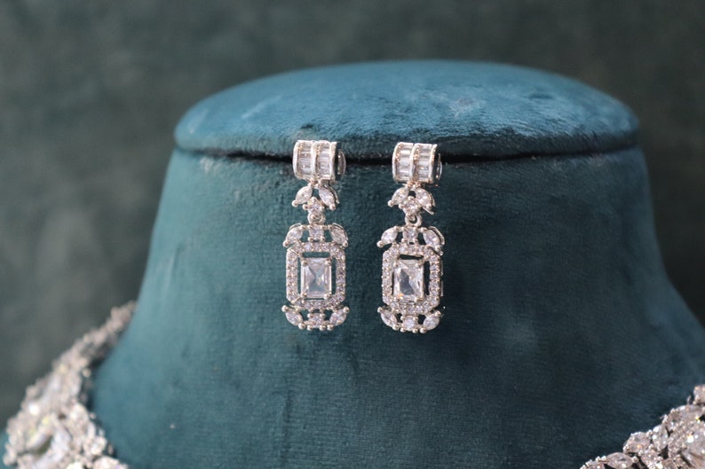 May include: Elegant silver-toned earrings with a rectangular, clear gemstone center and smaller surrounding stones. The earrings are displayed on a dark green velvet surface, showcasing their intricate design and sparkle. The earrings are part of a jewelry set.