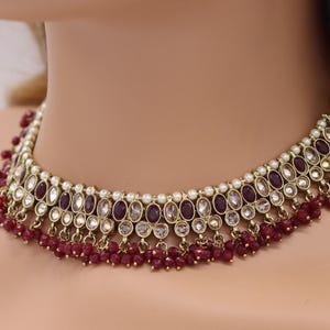 Puede incluir: Un collar de oro con una fila de piedras ovaladas en blanco y morado, con una franja de cuentas rojas.