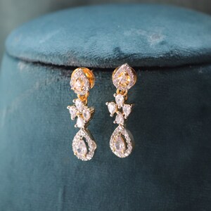 May include: Gold-toned drop earrings with clear, sparkling gemstones. The earrings feature a floral design with a teardrop-shaped stone at the bottom. The jewelry is displayed on a teal velvet surface.
