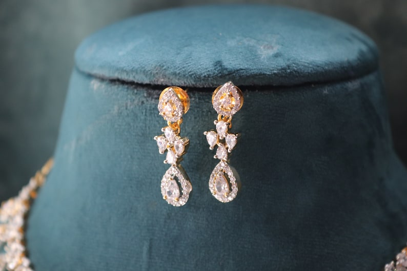 May include: Gold-toned drop earrings with clear, sparkling gemstones. The earrings feature a floral design with a teardrop-shaped stone at the bottom. The jewelry is displayed on a teal velvet surface.