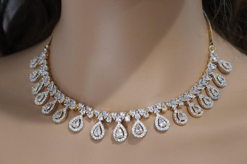 May include: A gold-toned necklace featuring a series of clear, teardrop-shaped pendants, each surrounded by smaller, sparkling stones. The necklace has a delicate, elegant design, perfect for special occasions.