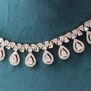 May include: Elegant necklace featuring a gold chain with teardrop-shaped pendants. Each pendant is encrusted with clear, sparkling gemstones, creating a luxurious and eye-catching design. The necklace is set against a teal background.