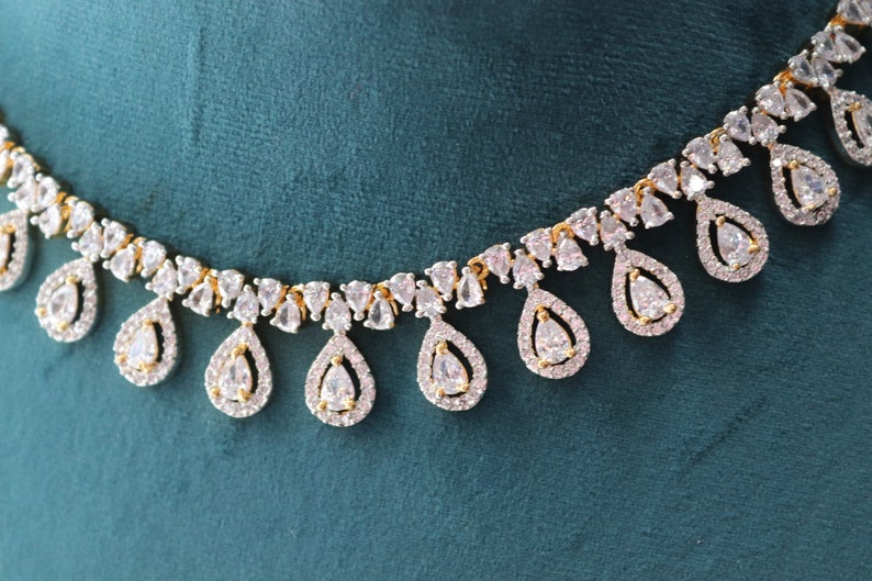 May include: Elegant necklace featuring a gold chain with teardrop-shaped pendants. Each pendant is encrusted with clear, sparkling gemstones, creating a luxurious and eye-catching design. The necklace is set against a teal background.
