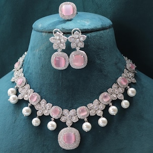Puede incluir: Un conjunto de joyas plateadas que incluye un collar, pendientes y un anillo. El collar y los pendientes tienen piedras centrales rosas rodeadas de cristales transparentes y detalles de perlas blancas. El anillo tiene una piedra rosa a juego.