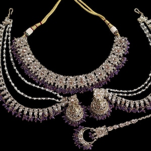 May include: A silver-toned bridal jewelry set with purple accents, including a necklace, earrings, and headpiece. The necklace and headpiece feature dangling strands and small purple beads. The earrings are round with a floral design.