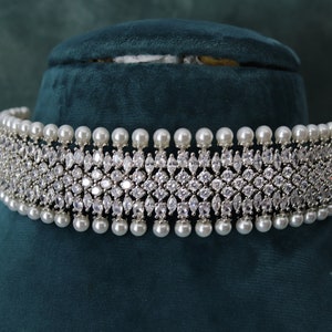 May include: A silver-toned choker necklace with a central band of sparkling, clear crystals in a geometric pattern. The necklace is accented with a row of white, round pearls along the top and bottom edges, and is displayed on a dark green velvet surface.
