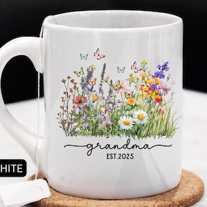 May include: White ceramic mug with a curved handle, featuring a floral design with butterflies and the word "grandma" with "EST.2025". The mug sits on a round cork coaster. A white tag is attached to the mug.